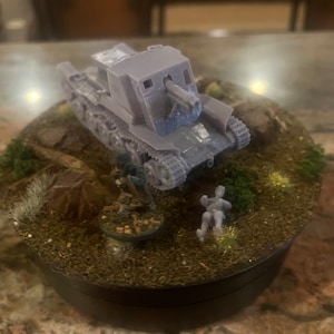 May include: A grey model tank with a long barrel is positioned on a miniature diorama base with small figures and terrain. The base is round with a dark rim. The tank is the main subject.