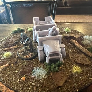 May include: A miniature diorama with a light grey truck and a soldier figurine. The scene is set on a circular base with a terrain of brown earth, green foliage, and small wooden logs. The truck has a detailed design.