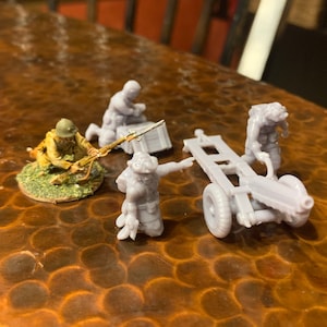 May include: Miniature military figures and a cannon. One figure is in a tan uniform, kneeling with a rifle. Other figures are in grey, with one operating a small cannon. The cannon has two wheels and a rectangular frame.