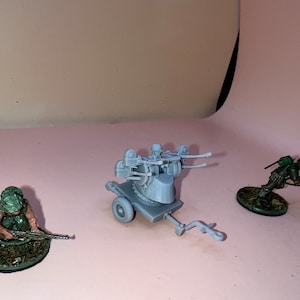 Op de afbeelding: Miniatuur militaire modellen op een roze achtergrond. De scène omvat een grijs luchtafweergeschut op een wagen en twee soldaten in groene uniformen. Eén soldaat ligt plat, de ander knielt.