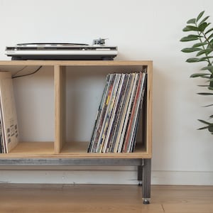 Peut inclure: Un meuble en bois pour tourne-disque avec trois étagères, une platine sur le dessus et une collection de disques vinyles. Le meuble a une base en métal et est posé sur un sol en bois.
