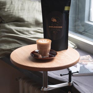 Könnte beinhalten: Ein kleiner, runder Holztisch, der an einem Heizkörper befestigt ist und eine Tasse Kaffee mit Latte Art auf einer Untertasse sowie einen dunklen Beutel mit Kaffeebohnen trägt. Der Beutel trägt den Text "GOLDDIEB" und ein Dachs-Logo.