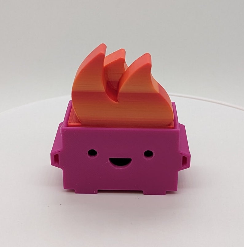 May include: A small, pink, 3D printed toy shaped like a brick with a smiling face and a bright orange and yellow flame on top.