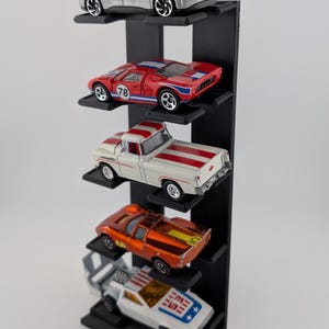May include: A vertical black display stand with five horizontal shelves holding five miniature toy cars. The cars include a silver sedan, a red race car with the number 78, a white pickup truck with red stripes, an orange sports car, and a white car.