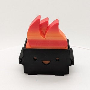 May include: A small black figurine with a smiling face and a red and orange flame on top. The figurine is made of plastic and has a cartoonish style.