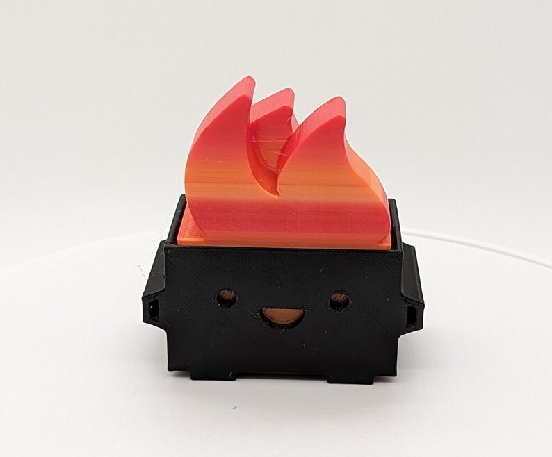 May include: A small black figurine with a smiling face and a red and orange flame on top. The figurine is made of plastic and has a cartoonish style.