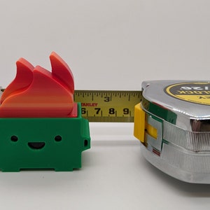 May include: A green 3D printed toy shaped like a trash can with a smiling face and a red, orange, and yellow flame on top. The toy is being measured with a tape measure that reads 8m/26'.