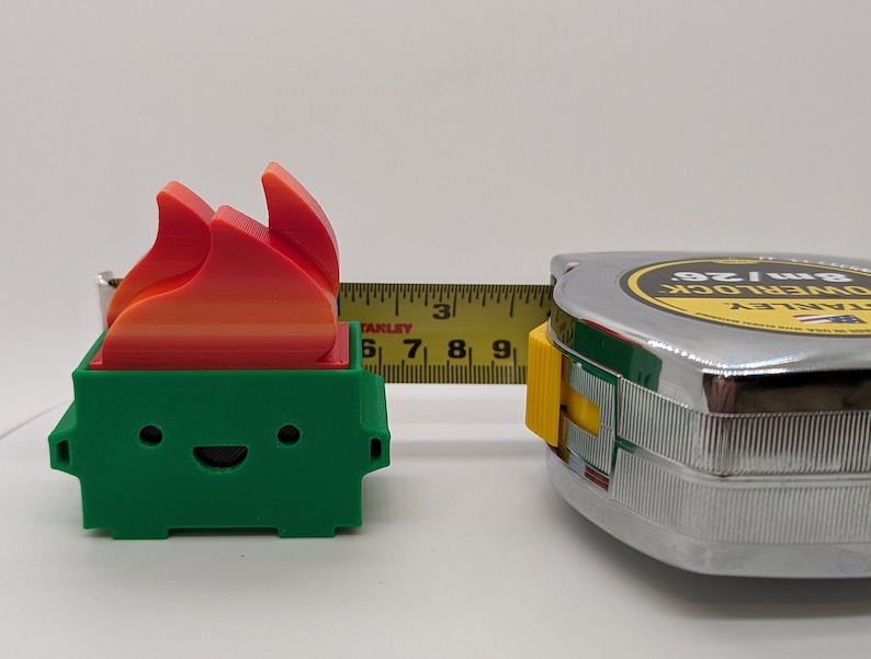 May include: A green 3D printed toy shaped like a trash can with a smiling face and a red, orange, and yellow flame on top. The toy is being measured with a tape measure that reads 8m/26'.