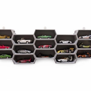 May include: A gray, honeycomb-shaped wall shelf displaying a collection of miniature toy cars. The cars are various colors, including yellow, red, green, and silver. The shelf is mounted on a white wall, creating a clean and organized display.