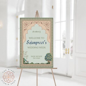 May include: A wedding welcome sign displayed on an easel. The sign features a light green backdrop with decorative tan and green borders. Text reads "Welcome to Sajanpreet's Wedding Week". A peacock and tree are illustrated at the base.