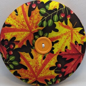 May include: A round, orange button with a white thread in the center, surrounded by a fabric circle with a pattern of yellow, orange, red, and black maple leaves.