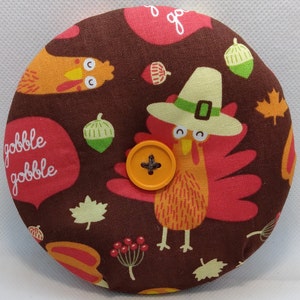 May include: A brown fabric Thanksgiving-themed round pillow with a turkey design. The turkey is wearing a pilgrim hat and has the words "gobble gobble" on the pillow.