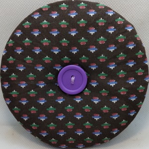 May include: A round, brown pin cushion with a repeating pattern of red, green, and blue shapes. A large, purple button is centered on the cushion. The cushion is likely used for sewing or crafting.