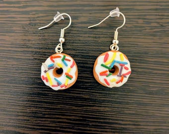 May include: A pair of donut earrings. Each earring features a miniature donut with white frosting and colorful sprinkles. The donuts have a brown base and a hole in the center. The earrings hang from silver-colored fish hook ear wires.