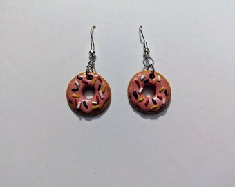 May include: A pair of pink donut earrings with colorful sprinkles. The donuts are made of polymer clay and have a silver hook.
