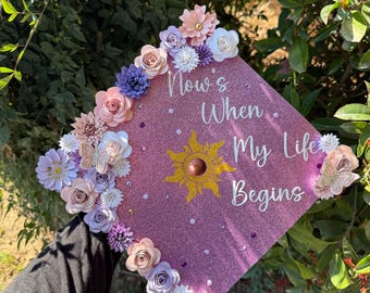 May include: A pink graduation cap decorated with paper flowers and a gold sun. The cap has the text "Now's When My Life Begins" written on it.
