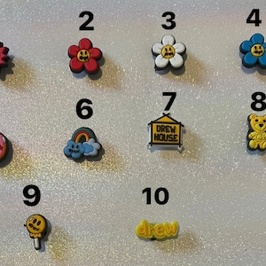May include: A selection of colourful shoe charms with diverse designs. These include a smiling sun, flowers, a rainbow, a donut with "DREW HOUSE" text, a house sign with "DREW HOUSE" text, a teddy bear, a lollipop, and the word "drew".
