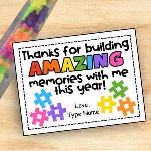 May include: A white card with the text "Thanks for building AMAZING memories with me this year!" in colorful letters. The card has colorful hashtag shapes and space to add a name. A clear tube filled with colorful building blocks is in the background.