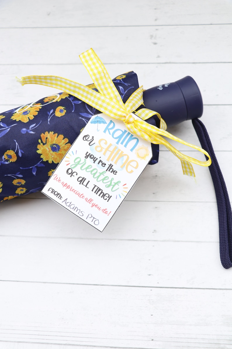Op de afbeelding: Een blauwe paraplu met een geel en wit geruit lint eromheen gebonden. Een wit cadeau-etiket met de tekst "Rain or Shine you're the greatest of all time! We appreciate all you do! From: Adams PTO" is aan het lint bevestigd.