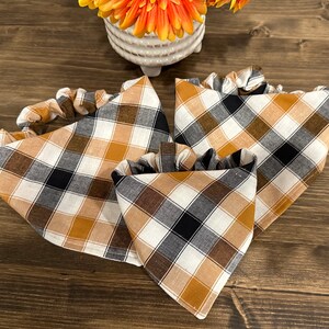 May include: A set of three plaid bandanas and a matching scrunchie in a fall color palette of brown, black, white, and gold. The bandanas are triangular, and the scrunchie is gathered.
