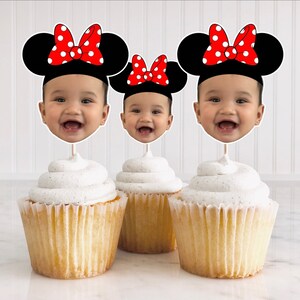 May include: Three cupcakes with white frosting topped with red and black Minnie Mouse themed cupcake toppers featuring a child's face.