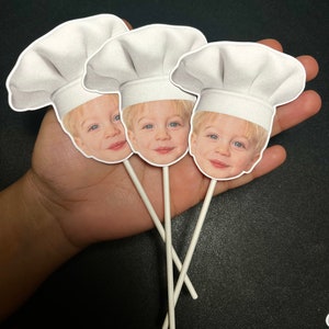 May include: Three white cupcake toppers with a child's face wearing a white chef's hat. The toppers are on white sticks.