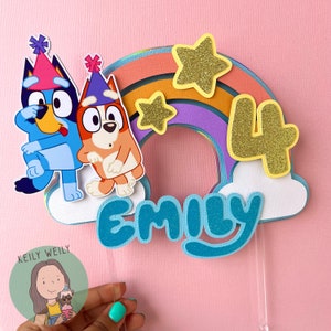 Peut inclure: Un topper de g&acirc;teau en forme de chien de dessin anim&eacute; bleu et orange avec un arc-en-ciel et des &eacute;toiles. Le topper de g&acirc;teau dit "Emily" et "4" en paillettes dor&eacute;es.