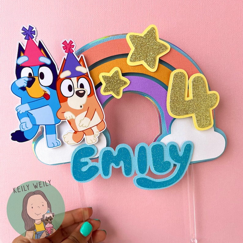 Peut inclure: Un topper de g&acirc;teau en forme de chien de dessin anim&eacute; bleu et orange avec un arc-en-ciel et des &eacute;toiles. Le topper de g&acirc;teau dit "Emily" et "4" en paillettes dor&eacute;es.