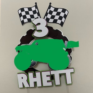 Puede incluir: Un adorno para pastel de camión monstruo verde con una bandera a cuadros, el número 3 y el nombre "Rhett" en letras blancas con un contorno morado.