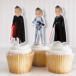 May include: Three cupcake toppers featuring Star Wars characters. The toppers are printed on white cardstock and feature a red lightsaber, a Stormtrooper, and Darth Vader. Each topper has a space for a photo to be inserted.