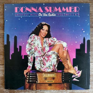 May include: Donna Summer Greatest Hits On The Radio Volumes I & II album cover featuring a woman in a floral dress sitting on a vintage radio against a city skyline at night.