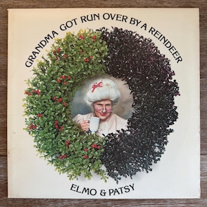May include: A book cover with the title "Grandma Got Run Over By A Reindeer" and the authors "Elmo & Patsy." The cover features a portrait of a woman inside a wreath of green and dark purple foliage with red berries. The woman is holding a mug.