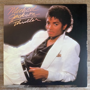 May include: Album cover featuring Michael Jackson in a white suit, black shirt, and black pants. The album title "Thriller" is written in a stylized font. The background is dark, and the image has a vintage aesthetic.