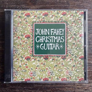 May include: A CD case featuring the album "John Fahey Christmas Guitar." The cover has a green and red floral pattern with a green rectangle containing the album title and artist name. The CD case is in a clear plastic case.