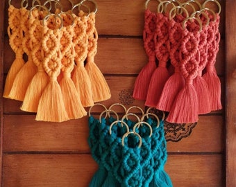 May include: A collection of macrame keychains featuring knotted cord designs in orange, red, and teal. Each keychain is attached to a gold-toned metal ring and finished with a long, straight tassel at the bottom, creating a textured appearance.