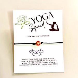 May include: A white card with the words "YOGA Squad" and a silhouette of a person in a yoga pose. A black cord bracelet with a red heart charm is attached to the card. The card includes the text "YOUR CUSTOM TEXT HERE".