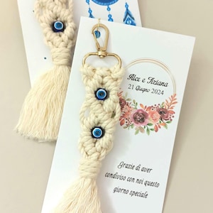 Puede incluir: Dos llaveros de macramé color hueso con cierres de mosquetón dorados, cada uno con dos cuentas de ojo turco azul y blanco. Los llaveros están sujetos a tarjetas blancas impresas con gráficos florales, el texto Alex e Tiziana 21 Giugno 2024 y Grazie di aver condiviso con noi questo giorno speciale.