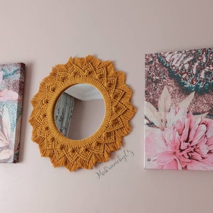 May include: A mustard yellow macrame mirror with a round mirror in the center. The mirror is hanging on a wall with two floral canvas prints on either side.