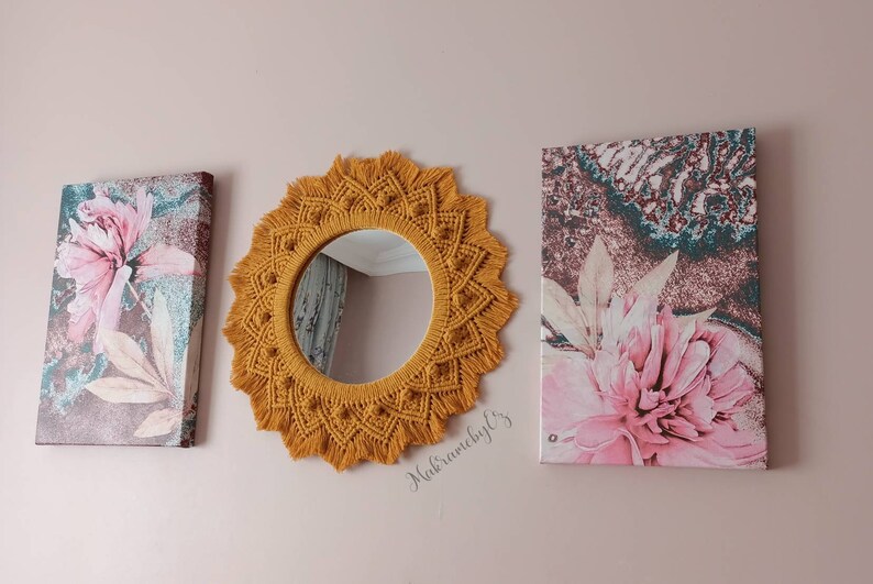May include: A mustard yellow macrame mirror with a round mirror in the center. The mirror is hanging on a wall with two floral canvas prints on either side.