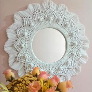 May include: White macrame mirror with a circular mirror in the center and a fringe border. The mirror is hanging on a pink wall with a bouquet of yellow and pink roses in the foreground.