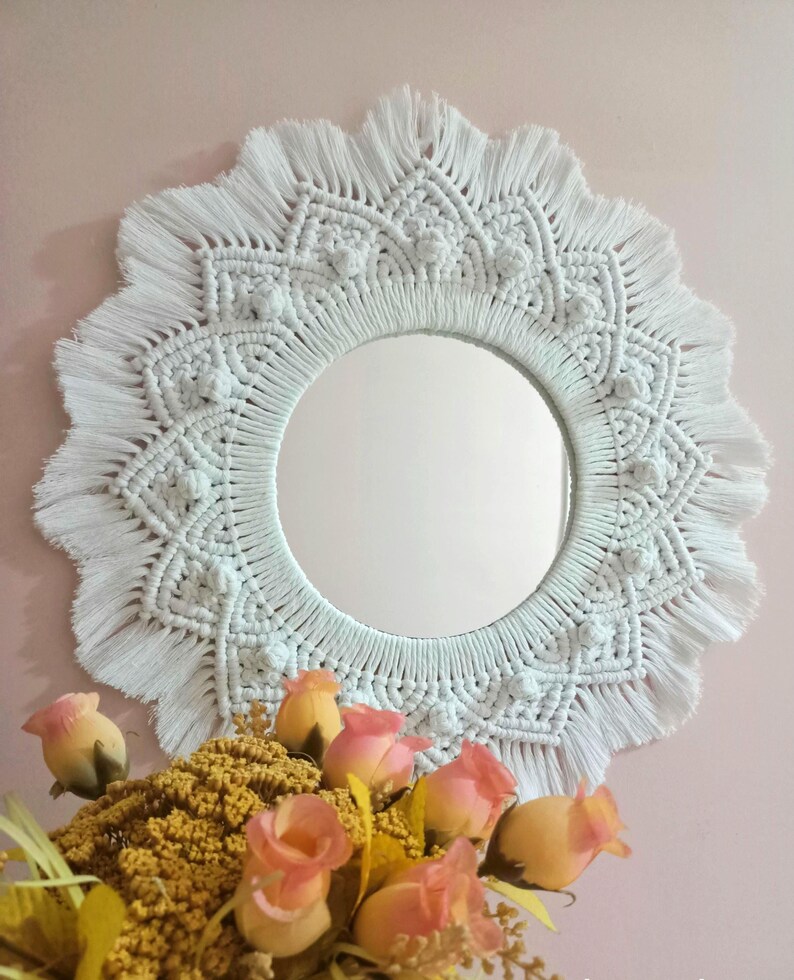 May include: White macrame mirror with a circular mirror in the center and a fringe border. The mirror is hanging on a pink wall with a bouquet of yellow and pink roses in the foreground.