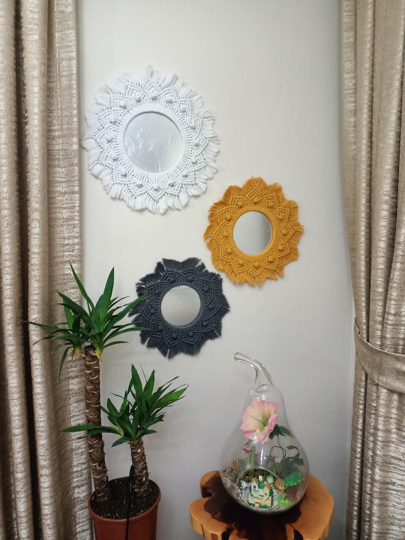 May include: Three macrame wall mirrors with different colored yarn. The mirrors are round and have a fringe border. The mirrors are hanging on a white wall.
