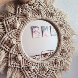 May include: A round macrame mirror with a beige color and a fringe border. The mirror is decorated with a geometric pattern and has a knot detail around the edge.