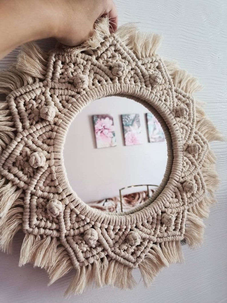 May include: A round macrame mirror with a beige color and a fringe border. The mirror is decorated with a geometric pattern and has a knot detail around the edge.