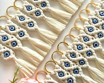 May include: A collection of macrame keychains made from off-white cotton cord, each featuring a blue and white evil eye bead and a matching tassel. The keychains are attached to gold-toned metal rings and arranged in a diagonal pattern.