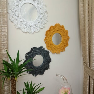 May include: Three macrame wall mirrors with round mirrors in the center. The mirrors are white, yellow, and gray. The mirrors are hanging on a white wall.