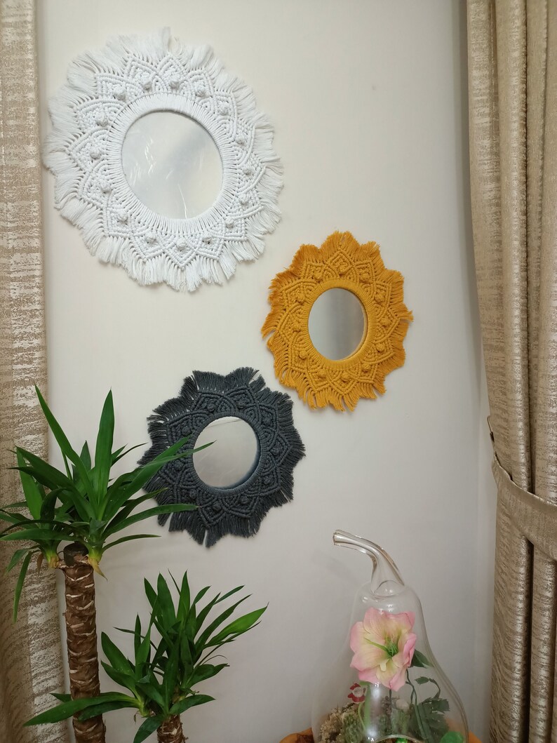 May include: Three macrame wall mirrors with round mirrors in the center. The mirrors are white, yellow, and gray. The mirrors are hanging on a white wall.