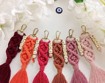 Pode incluir: Uma coleção de chaveiros de macramê feitos à mão em várias cores, incluindo bordô, laranja, rosa e marrom. Cada chaveiro apresenta uma borla e contas de madeira com nomes. Fechos dourados são anexados no topo.