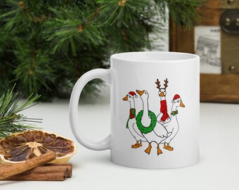 Op de afbeelding: Witte keramische mok met een feestelijk ontwerp van vier ganzen. De ganzen zijn versierd met kerstaccessoires, waaronder kerstmutsen, een krans en een rendiergeweige haarband. De mok heeft een gebogen handvat.