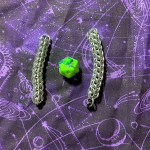 May include: Two silver chainmail bracelets on a purple background with white stars and a green and yellow dice with a number 8 on it.